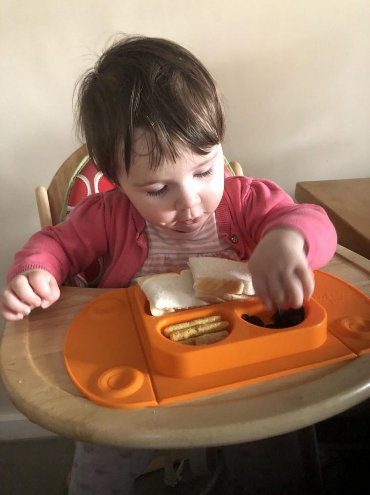 Lottie eating her lunch from an orange Mini EasyMat. Suctioned to a wooden high chair tray